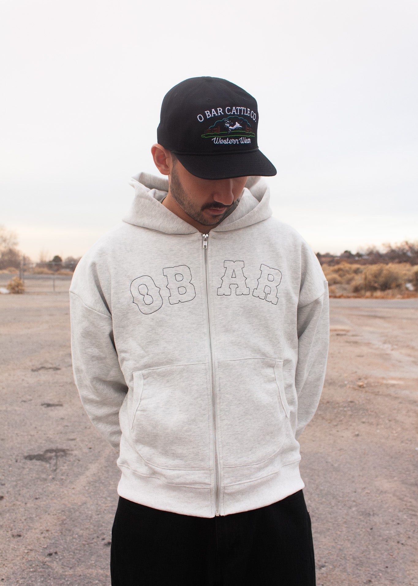 Person wearing a gray hoodie and black cap with 'O Bar Cattle Co.' branding in an open area.